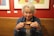 An older woman with white hair enjoys a hot beverage in a ceramic mug. She is wearing a denim jacket, and there are colorful paintings on a red wall in the background. She appears content and relaxed, sitting in a cozy café environment.