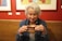 An older woman with white hair enjoys a hot beverage in a ceramic mug. She is wearing a denim jacket, and there are colorful paintings on a red wall in the background. She appears content and relaxed, sitting in a cozy café environment.