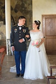 A couple stands indoors, with the man wearing a military uniform and the woman wearing a white wedding dress. They are facing each other, smiling, and the woman is holding a bouquet of pink and white roses. The background features a stone wall and a wooden door.