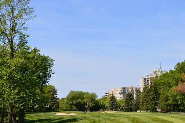 A lush golf course with well-maintained grass and sand bunkers. Tall green trees border the course, and in the distance, there are several modern buildings partially obscured by trees. The sky above is clear and blue.
