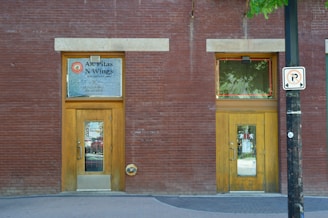 A brick building facade featuring two wooden doors with glass panes and signs. The left door has a sign for 'AK Pitas N Wings' above it, and there's a 'no parking' sign attached to a black post in front of the right door. The sidewalk in front of the building is concrete, and there are green leaves from a tree partially visible at the top right corner.