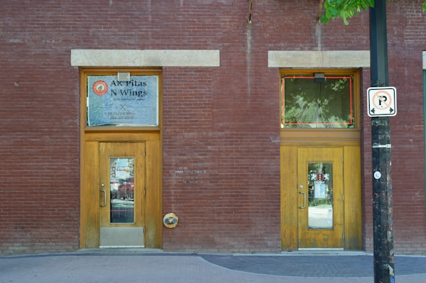 A brick building facade featuring two wooden doors with glass panes and signs. The left door has a sign for 'AK Pitas N Wings' above it, and there's a 'no parking' sign attached to a black post in front of the right door. The sidewalk in front of the building is concrete, and there are green leaves from a tree partially visible at the top right corner.