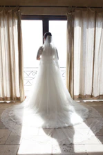A bride gently holding the flowing train of her classic white wedding dress in a sunlit room.