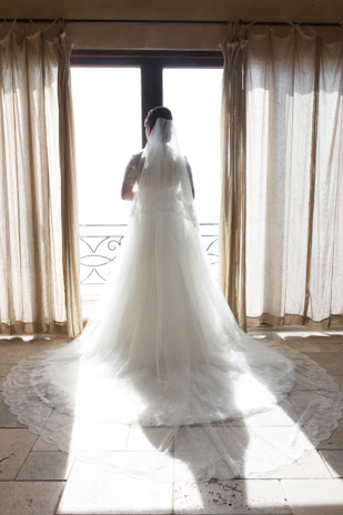 A bride gently holding the flowing train of her classic white wedding dress in a sunlit room.