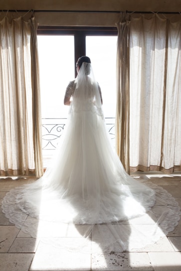 A bride smiling softly as a stylist adjusts her veil in a sunlit room.