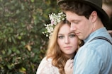 A couple stands closely together in a natural setting. The woman, adorned with a floral headpiece, looks directly into the camera while the man, wearing a hat, gazes downwards. The background is filled with lush greenery, enhancing the intimate and serene atmosphere.