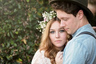 A couple stands closely together in a natural setting. The woman, adorned with a floral headpiece, looks directly into the camera while the man, wearing a hat, gazes downwards. The background is filled with lush greenery, enhancing the intimate and serene atmosphere.