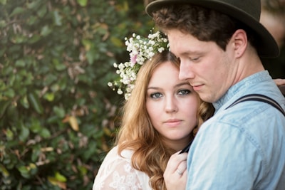 A couple stands closely together in a natural setting. The woman, adorned with a floral headpiece, looks directly into the camera while the man, wearing a hat, gazes downwards. The background is filled with lush greenery, enhancing the intimate and serene atmosphere.