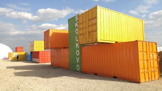 Several colorful shipping containers are stacked on a gravel surface. The containers are painted in bright colors, including yellow, orange, green, blue, and red. A person is visible walking past the containers. The word 'SKOLKOVO' is painted on some of the containers. The sky is clear with a few clouds, suggesting a sunny day.
