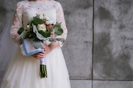 A bride in an elegant lace wedding dress is holding a bouquet of flowers. The flowers include roses and eucalyptus leaves, wrapped in a white and blue cloth. The background is a textured gray wall.