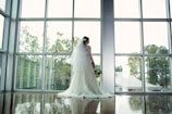 A bride, wearing a white wedding gown with a long veil, stands in front of large windows. She holds a bouquet of flowers and looks out at the landscape outside, which includes trees and a building with a metal roof. Soft light comes through the windows, casting reflections on the polished floor.