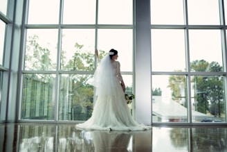 A bride, wearing a white wedding gown with a long veil, stands in front of large windows. She holds a bouquet of flowers and looks out at the landscape outside, which includes trees and a building with a metal roof. Soft light comes through the windows, casting reflections on the polished floor.