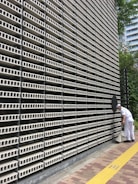 A large, repetitive pattern of rectangular concrete blocks forms the facade of a modern building, creating a grid-like appearance. A person dressed in white work attire is engaged in cleaning, using a long-handled dustpan and broom on the sidewalk adjacent to the structure. A yellow tactile paving strip runs along the path, providing guidance for visually impaired pedestrians. Trees and another building are visible in the background.