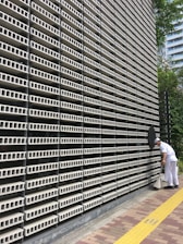 A large, repetitive pattern of rectangular concrete blocks forms the facade of a modern building, creating a grid-like appearance. A person dressed in white work attire is engaged in cleaning, using a long-handled dustpan and broom on the sidewalk adjacent to the structure. A yellow tactile paving strip runs along the path, providing guidance for visually impaired pedestrians. Trees and another building are visible in the background.
