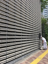 A large, repetitive pattern of rectangular concrete blocks forms the facade of a modern building, creating a grid-like appearance. A person dressed in white work attire is engaged in cleaning, using a long-handled dustpan and broom on the sidewalk adjacent to the structure. A yellow tactile paving strip runs along the path, providing guidance for visually impaired pedestrians. Trees and another building are visible in the background.