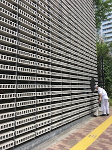 A large, repetitive pattern of rectangular concrete blocks forms the facade of a modern building, creating a grid-like appearance. A person dressed in white work attire is engaged in cleaning, using a long-handled dustpan and broom on the sidewalk adjacent to the structure. A yellow tactile paving strip runs along the path, providing guidance for visually impaired pedestrians. Trees and another building are visible in the background.