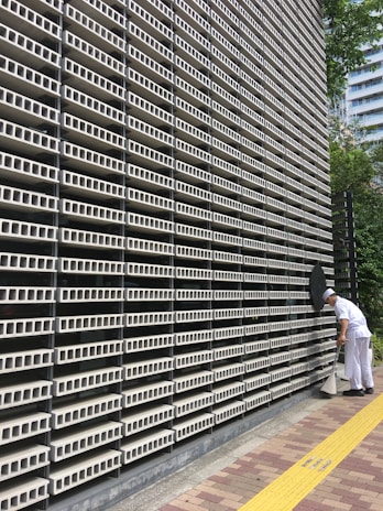 A large, repetitive pattern of rectangular concrete blocks forms the facade of a modern building, creating a grid-like appearance. A person dressed in white work attire is engaged in cleaning, using a long-handled dustpan and broom on the sidewalk adjacent to the structure. A yellow tactile paving strip runs along the path, providing guidance for visually impaired pedestrians. Trees and another building are visible in the background.