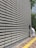 A large, repetitive pattern of rectangular concrete blocks forms the facade of a modern building, creating a grid-like appearance. A person dressed in white work attire is engaged in cleaning, using a long-handled dustpan and broom on the sidewalk adjacent to the structure. A yellow tactile paving strip runs along the path, providing guidance for visually impaired pedestrians. Trees and another building are visible in the background.