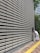 A large, repetitive pattern of rectangular concrete blocks forms the facade of a modern building, creating a grid-like appearance. A person dressed in white work attire is engaged in cleaning, using a long-handled dustpan and broom on the sidewalk adjacent to the structure. A yellow tactile paving strip runs along the path, providing guidance for visually impaired pedestrians. Trees and another building are visible in the background.