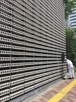 A large, repetitive pattern of rectangular concrete blocks forms the facade of a modern building, creating a grid-like appearance. A person dressed in white work attire is engaged in cleaning, using a long-handled dustpan and broom on the sidewalk adjacent to the structure. A yellow tactile paving strip runs along the path, providing guidance for visually impaired pedestrians. Trees and another building are visible in the background.