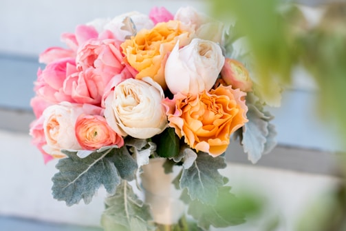 A bouquet of colorful, blooming flowers consisting of vibrant orange, pink, and cream roses, surrounded by lush, grey-green leaves. The flowers are tightly arranged and appear fresh and vibrant against a soft, blurred background.