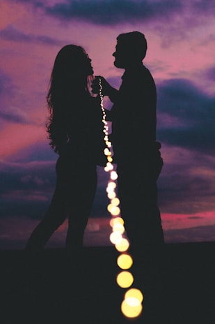 Silhouetted figures of a couple facing each other against a vibrant sunset. A string of lights runs from the foreground to their hands, creating a sense of connection and intimacy.