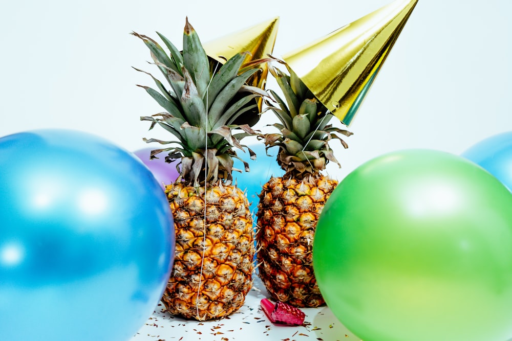 two pineapples with gold party hats near colorful balloons