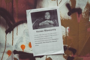 A printed paper with text and a black-and-white photograph of a woman is pasted against a wall covered in colorful graffiti. The text appears to be in Russian and includes details about an individual named Colleen McCullough, mentioning dates and places. The wall has abstract patterns in various colors, including brown, orange, and white.
