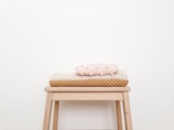 A minimalist and cozy scene with a small wooden stool against a plain off-white wall. Neatly folded knit blankets in pastel pink and beige tones are stacked on top of the stool.