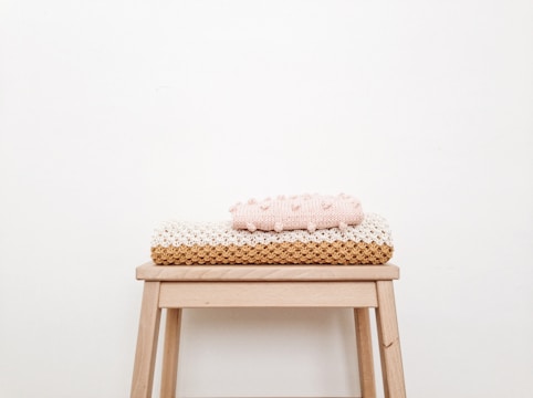 A minimalist and cozy scene with a small wooden stool against a plain off-white wall. Neatly folded knit blankets in pastel pink and beige tones are stacked on top of the stool.