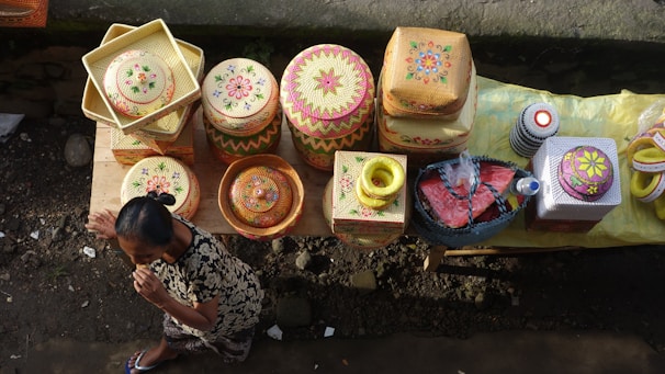 A person is arranging intricately designed baskets displayed on a wooden table. The baskets are woven and feature colorful floral patterns. The setting appears to be outdoors with a stone pavement backdrop.
