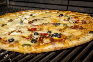 A pizza with a golden-brown crust is topped with melted cheese, black olives, green bell peppers, red bell peppers, and slices of pepperoni. The pizza appears to be cooking on a grill.