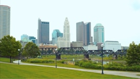 A vibrant urban skyline featuring modern skyscrapers with a variety of architectural designs. In the foreground, a lush green park with a winding pathway is bordered by neatly manicured trees. A bridge extends across a calm river, adding an industrial element to the serene park setting.