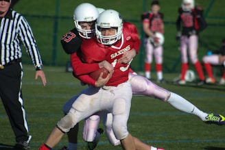 A dynamic scene from an American football game shows a player in a red jersey and white helmet attempting to move forward while holding the ball. He is being tackled by another player in a black jersey. A referee dressed in a black and white striped uniform is visible nearby, observing the play. Additional players and a grassy field can be seen in the background.