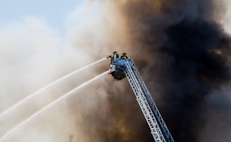 Two firefighters are positioned on an elevated platform at the top of a long extended fire truck ladder, directing powerful streams of water at a dense, dark cloud of smoke. The sky is partially visible, providing a stark contrast against the thick, swirling black and gray smoke.