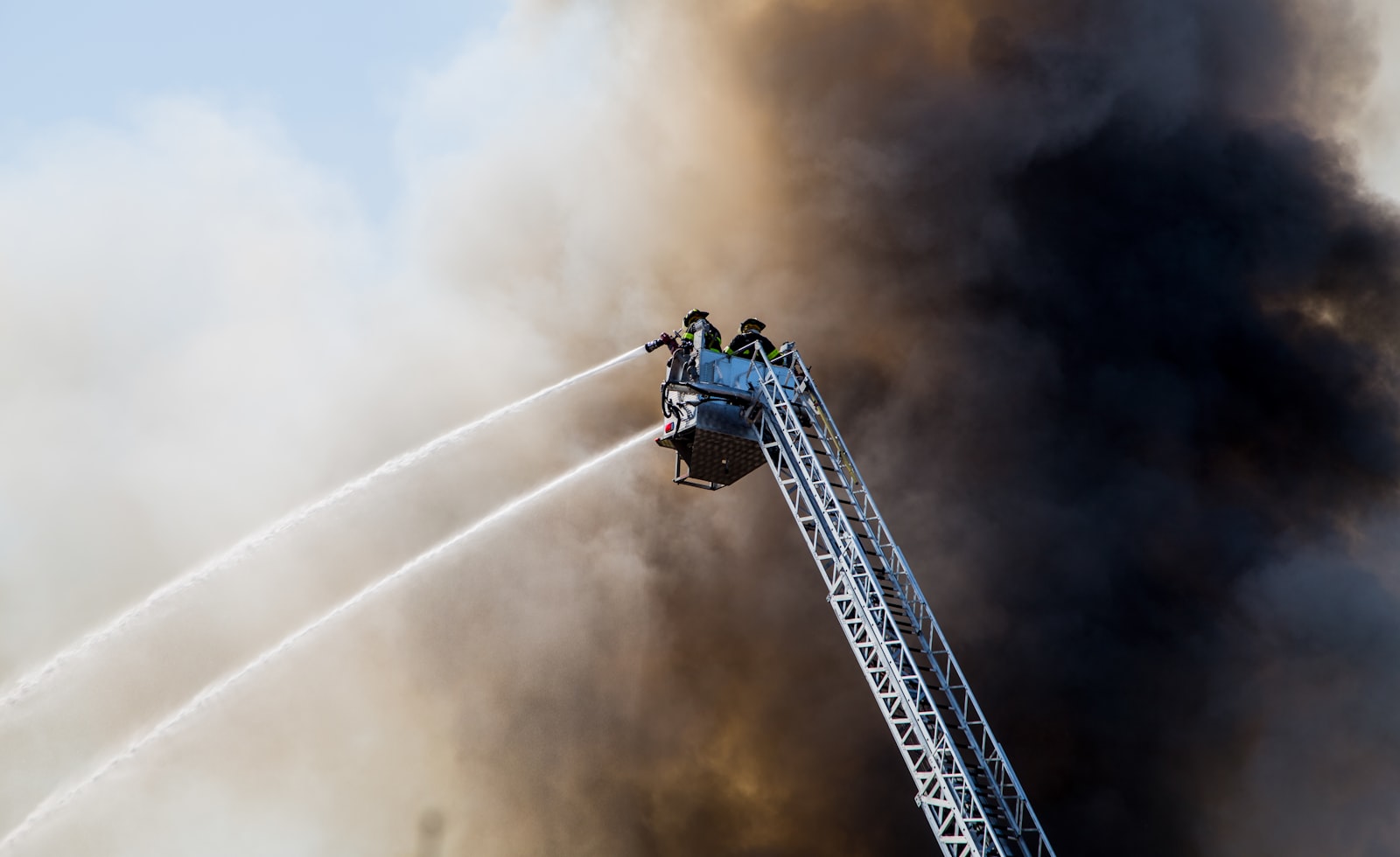 Firefighters on a ladder spraying water into smoke during an active fire scene