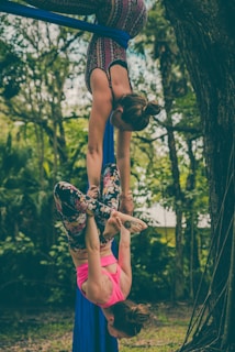 Two people are performing an aerial silk routine in an outdoor setting surrounded by trees and greenery. One person is hanging upside down from the silks while the other supports them with their legs wrapped around the fabric.