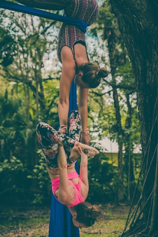 Two people are performing an aerial silk routine in an outdoor setting surrounded by trees and greenery. One person is hanging upside down from the silks while the other supports them with their legs wrapped around the fabric.