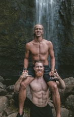 a man is holding a man on his shoulders in front of a waterfall