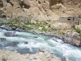 A flowing river with clear blue water winding through a rocky landscape, bordered by large boulders and patches of green grass. A small stone structure is nestled against the rocky hillside.
