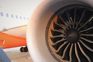 Close-up view of an airplane turbine engine, showcasing the intricate metal blades within the engine casing. The fuselage of the aircraft, painted in an orange and white color scheme, is visible in the background, along with part of the landing gear.
