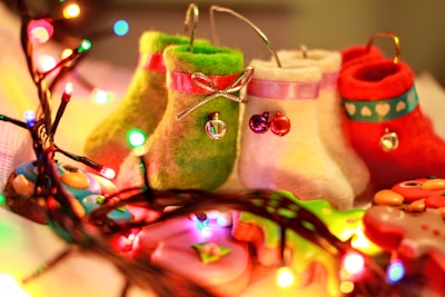 A collection of colorful, decorative felt stockings and ornaments surrounded by bright, multicolored Christmas lights. Each stocking is uniquely designed with ribbons and small bells, creating a festive and cheerful atmosphere.