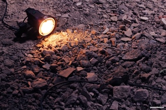 A portable spotlight illuminates a patch of rough, rocky ground. The light casts a warm glow over the uneven surface, highlighting the texture of the rubble and stones scattered around. An electrical cord is attached to the light, trailing off into the shadows.