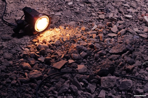 A portable spotlight illuminates a patch of rough, rocky ground. The light casts a warm glow over the uneven surface, highlighting the texture of the rubble and stones scattered around. An electrical cord is attached to the light, trailing off into the shadows.