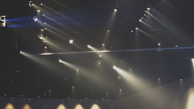 A dark theater or concert hall ceiling with numerous spotlights casting beams of light in various directions. The lights create a dramatic array of rays, some of which highlight particles in the air, contributing to the ambient atmosphere.