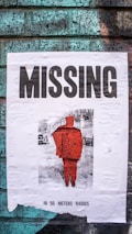 A weathered, torn poster is attached to a brick wall. The poster has the word 'MISSING' in large bold letters at the top. Below it, there is a faded image of a person in a bright red silhouette against a monochrome background. Text at the bottom reads 'IN 50 METERS RADIUS'. The brick wall has a weathered look with green and blue tones.