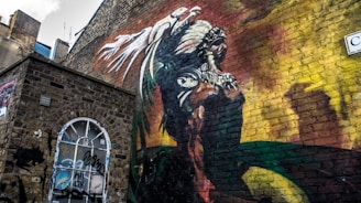 A brick wall features a vibrant and intricate mural depicting a figure wearing a detailed headdress, possibly resembling a mythical or tribal warrior. The artwork extends across the wall with bold colors and dynamic shapes. Below the mural, there is a boarded-up window covered with various graffiti and stickers.