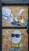 The artwork features a collage of comic book pages in the background with a prominent illustration of a large Spiderman-like mask, alongside a cartoonish figure wearing sunglasses and a balaclava, riding a bicycle and blowing a bubble. Smaller elements include parachuting creatures and an abstract figure holding a pencil.