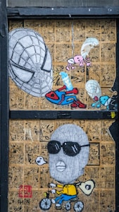 The artwork features a collage of comic book pages in the background with a prominent illustration of a large Spiderman-like mask, alongside a cartoonish figure wearing sunglasses and a balaclava, riding a bicycle and blowing a bubble. Smaller elements include parachuting creatures and an abstract figure holding a pencil.