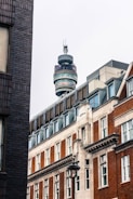 A cityscape featuring a tall telecommunications tower rising above a mix of modern and historic brick buildings. The architecture displays a combination of glass, steel, and traditional bricks with detailed window frames. The tower has multiple circular viewing platforms and antennas on top.